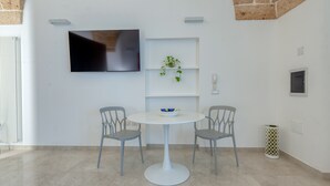 A modern room with a round white table, two grey chairs, a TV mounted on the wall, a small plant on a shelf, and a trash can.