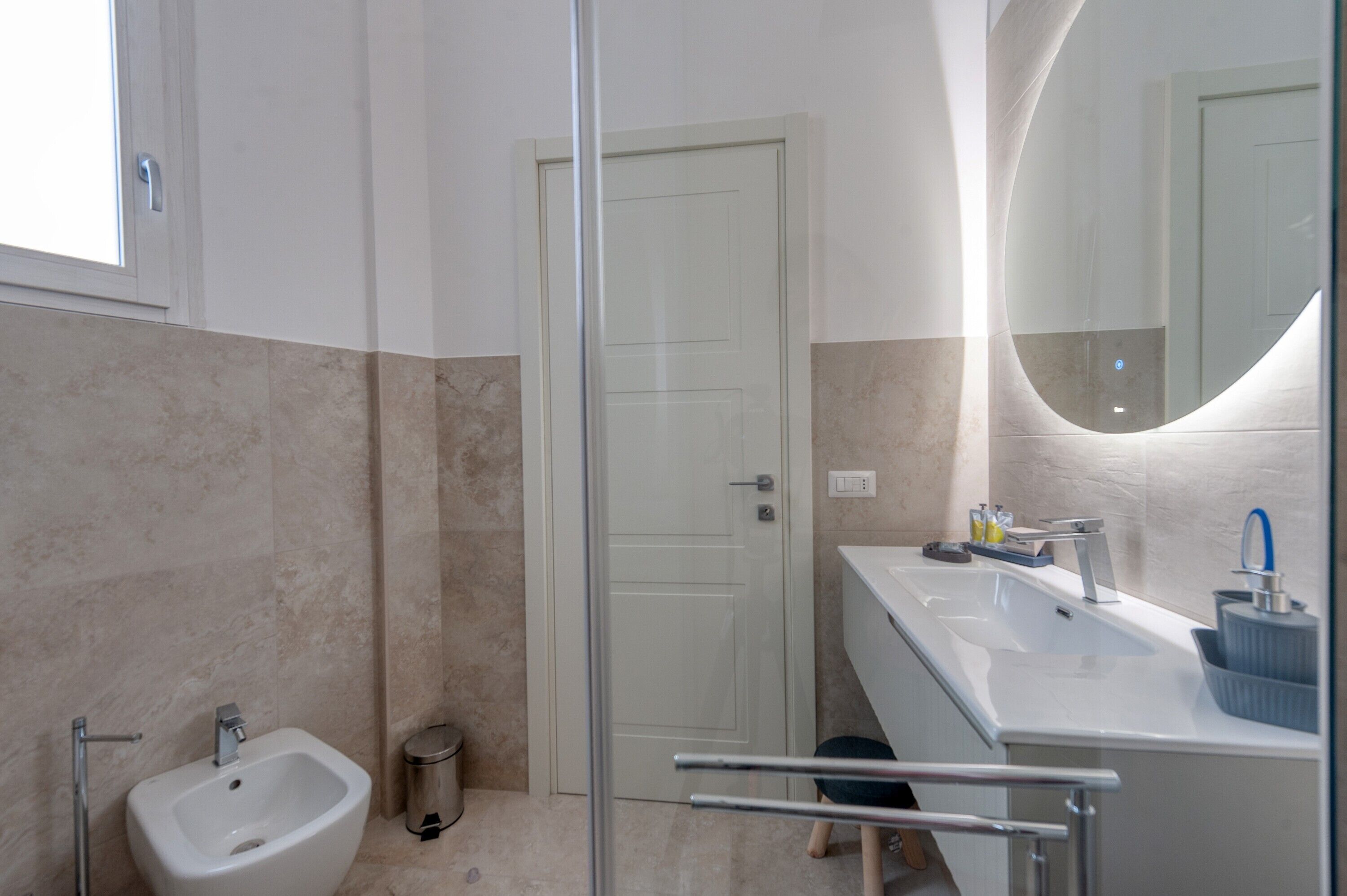 Une salle de bain moderne avec une douche à parois de verre, un lavabo blanc doté d’un robinet bleu et un miroir à cadre incurvé.