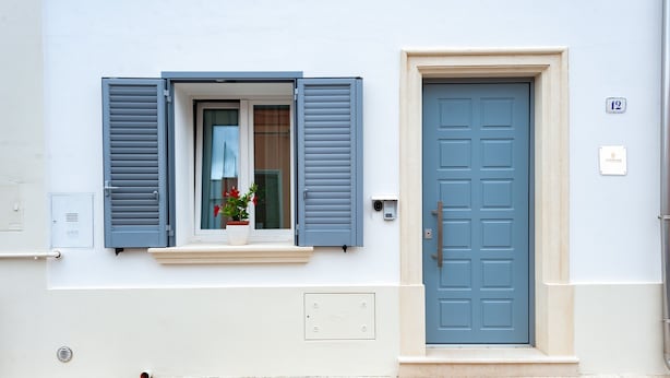 Eine Gebäudefassade mit einer blauen Tür, einem Fenster mit Rollläden und einem kleinen Blumentopf.