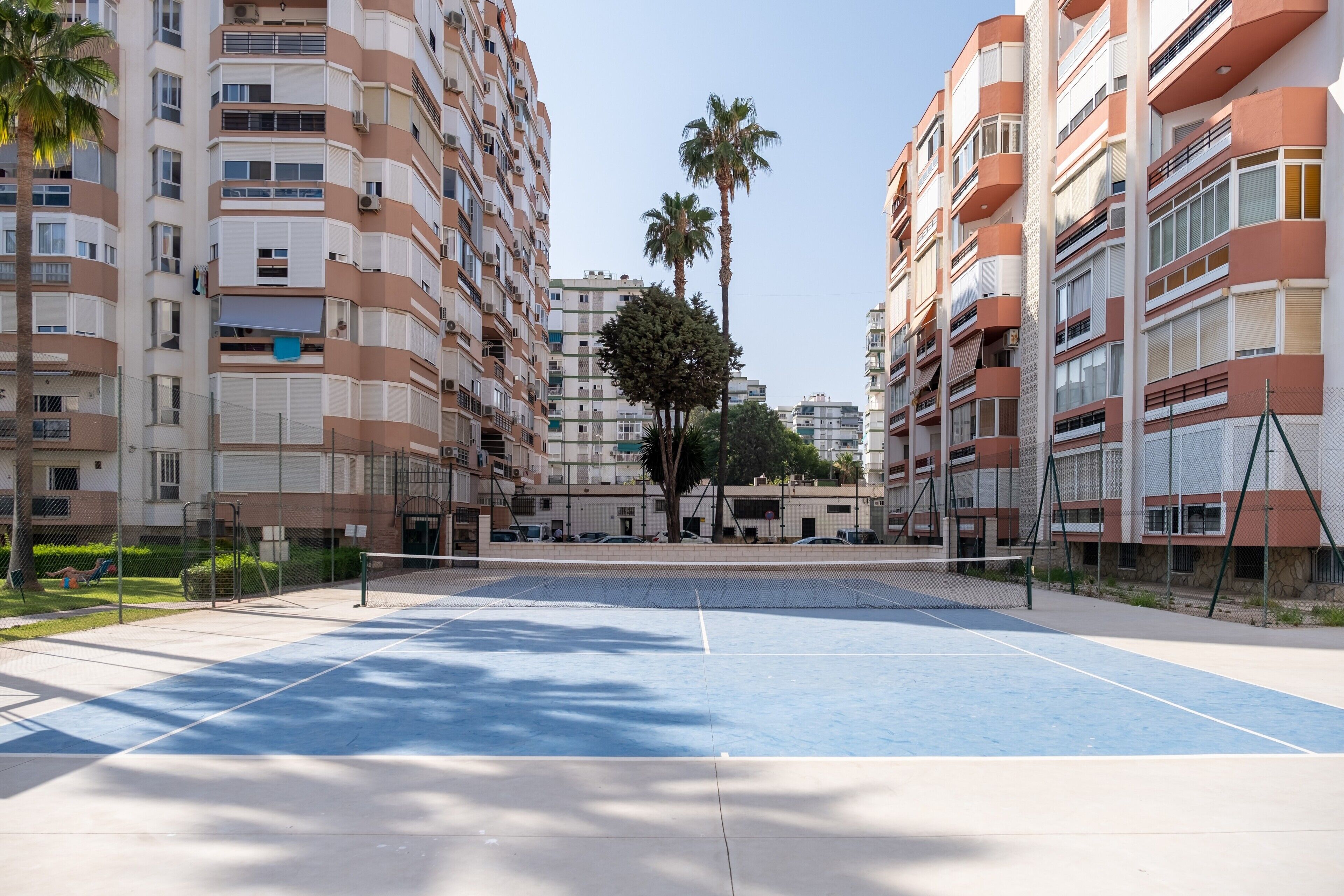 Un edificio residencial de varios pisos con balcones, cancha de tenis y palmeras.