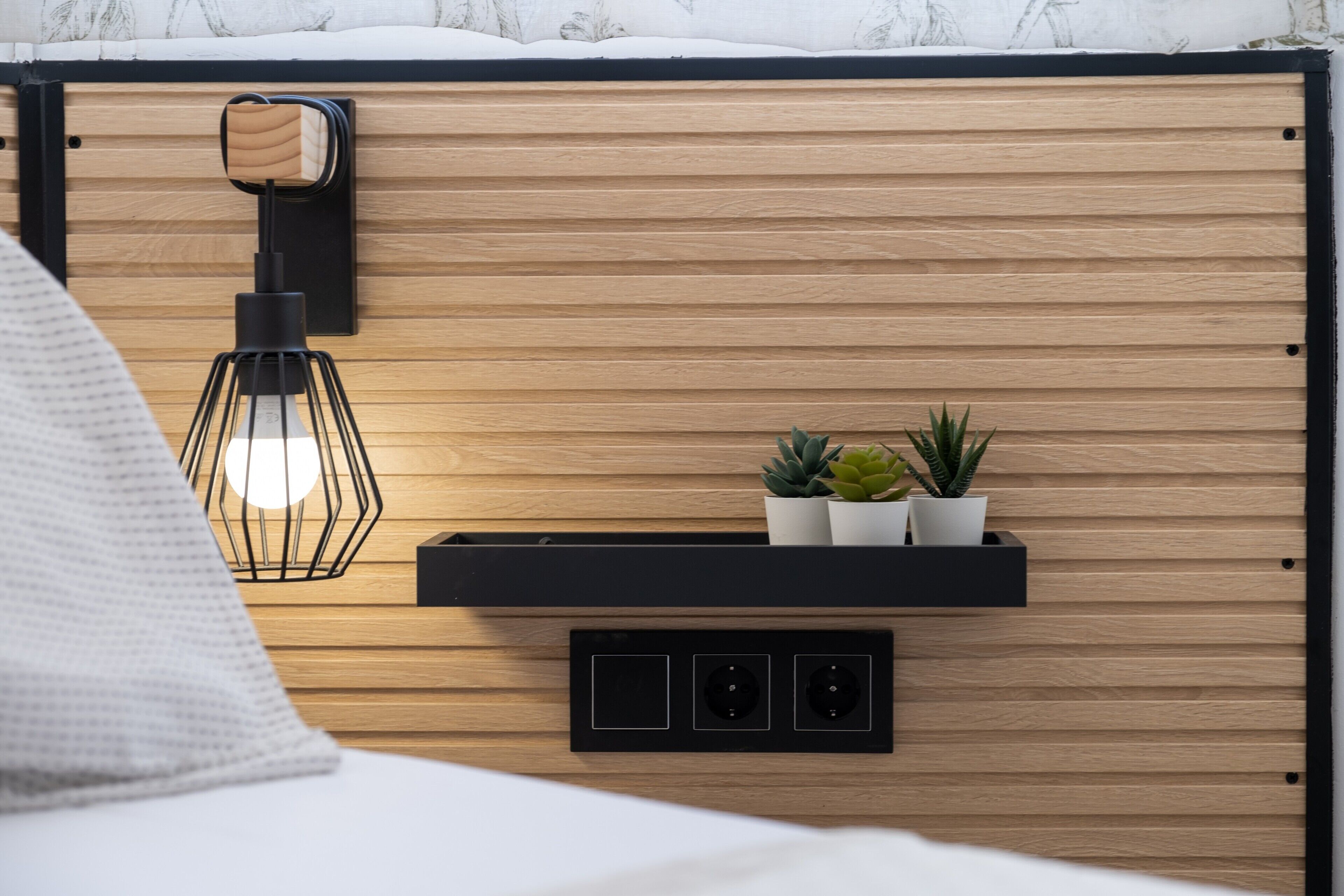 A bed with a white pillow, a bedside lamp, a wall-mounted shelf with two potted plants, and electrical outlets.