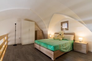 Une chambre avec un plafond en pente, un lit recouvert d’une couverture verte, une table de chevet avec une lampe, une armoire en bois et un ours en peluche sur le lit.