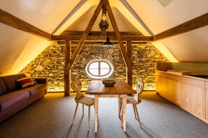 Une pièce mansardée chaleureuse avec un mur en pierre, des poutres apparentes, une fenêtre ronde, une table à manger et un canapé.