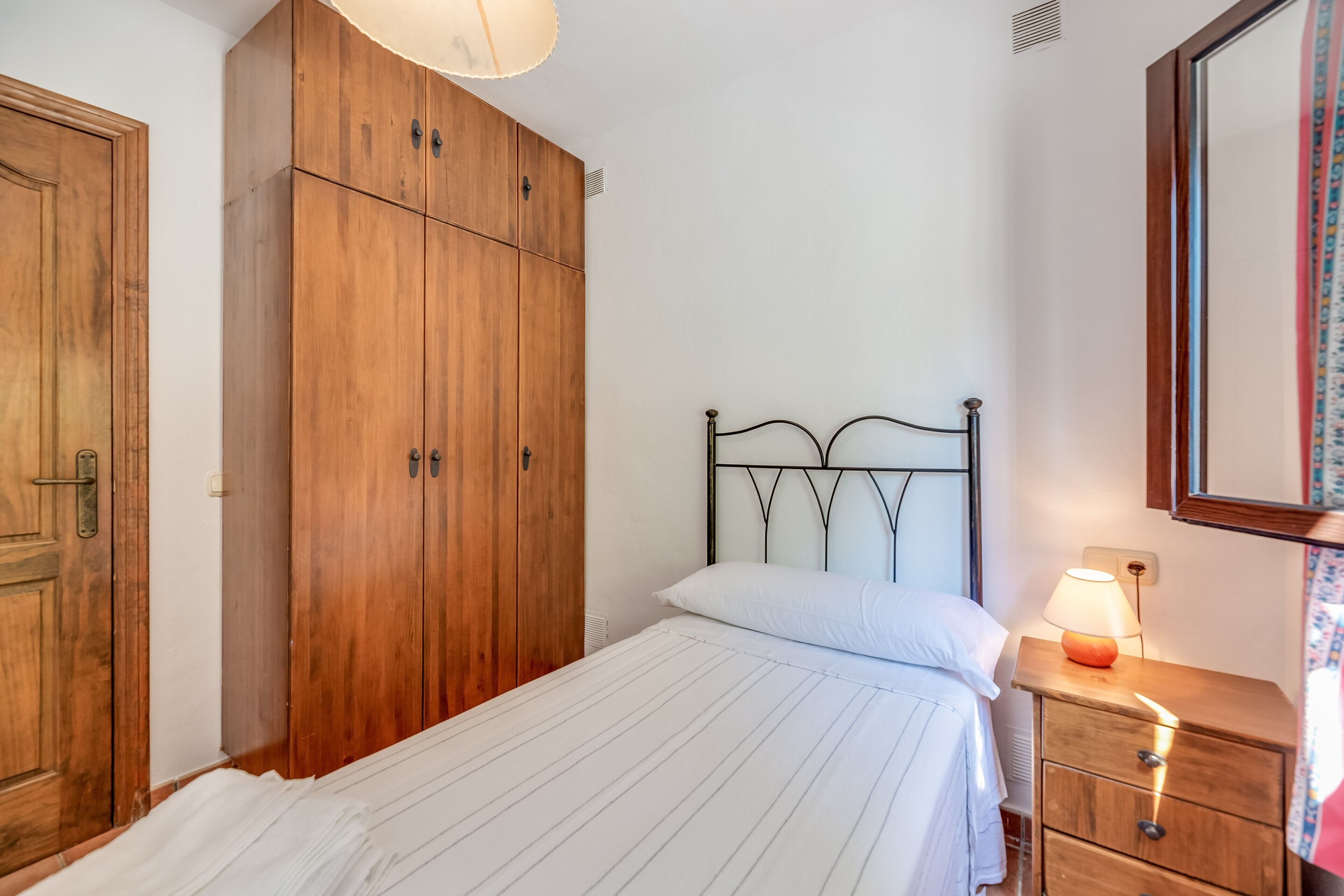 A bedroom with a bed, wooden wardrobe, bedside table, lamp, and a mirror.