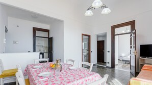 A dining area with a table set for four, white chairs, a kitchen in the background, and a TV in the corner.