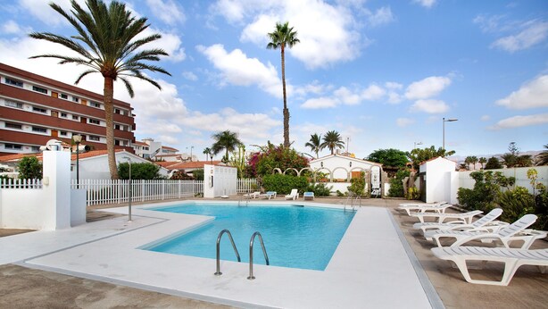 A swimming pool with lounge chairs, palm trees, and a hotel building in the background. - Vacation Home 'Haus Brigitte' with Shared Pool, Wi-Fi and Air Conditioning (Playa del Inglés)
