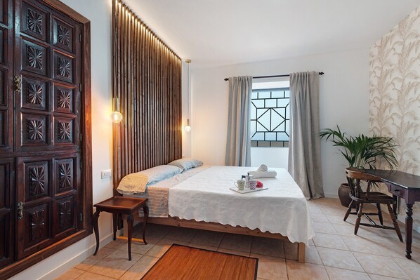 A bedroom with a bed, bedside table, lamp, window with curtains, and a door with intricate design.