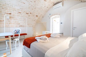 Een slaapkamer met een bakstenen plafond, een bed met witte lakens, een wit bureau met een stoel en een raam met uitzicht.