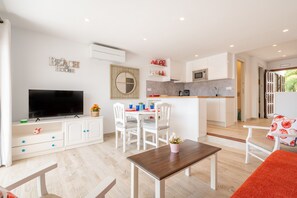 Un salon moderne doté d’un téléviseur à écran plat, d’un coin repas avec des chaises, d’une cuisine avec des meubles blancs et d’une grande fenêtre.