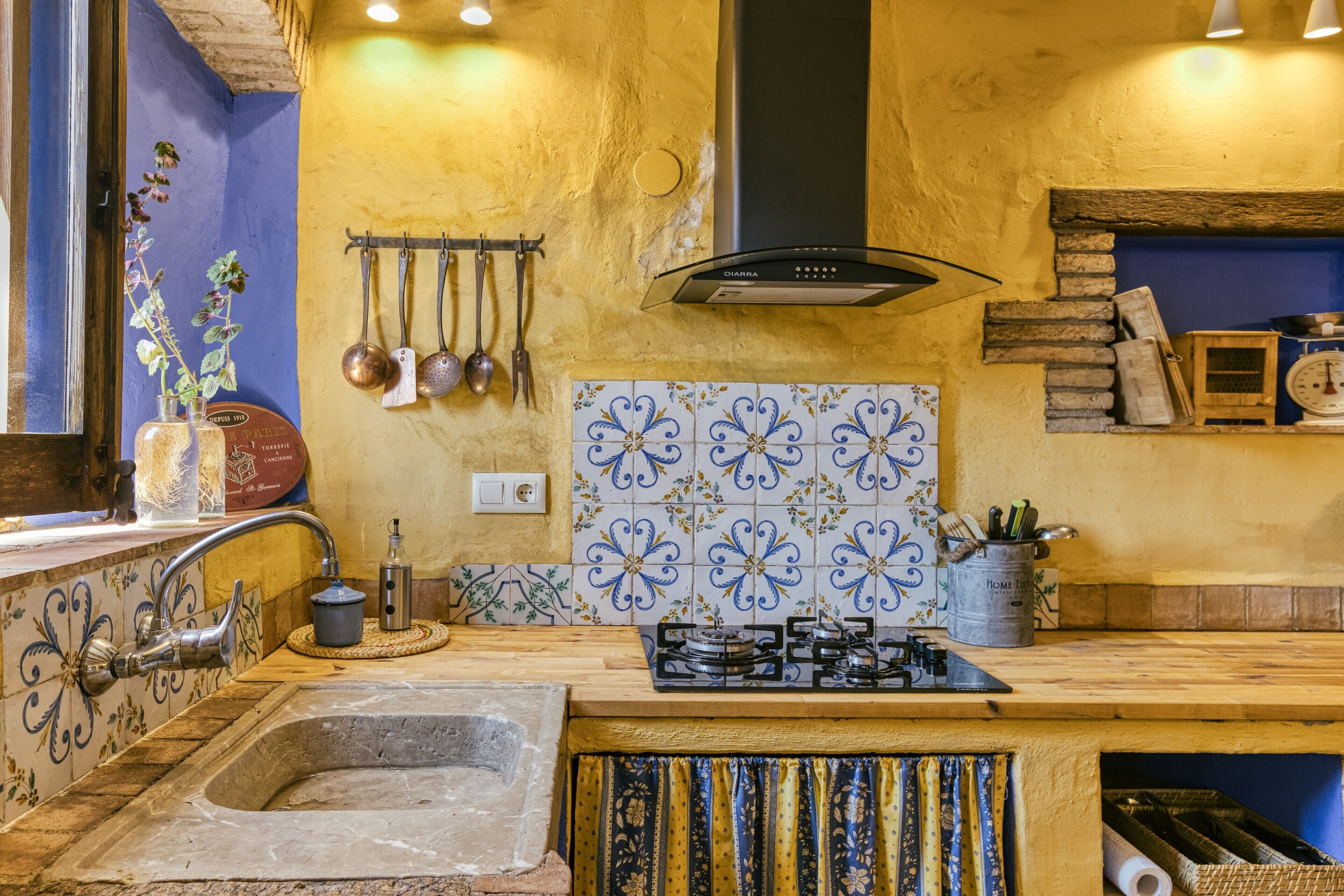 A kitchen with a stone sink, gas stove, hanging utensils, and decorative tiles.