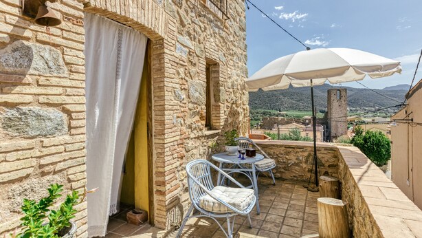 A stone building with a balcony, white curtains, wicker chairs, a table with a potted plant, and a white umbrella.