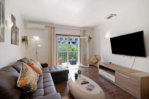 A modern living room with a sofa, ottoman, coffee table, TV, and a balcony with plants.