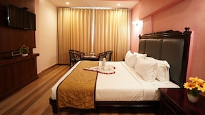A hotel room with a bed, a television, a ceiling fan, a window with curtains, a small table with a vase of flowers, and a wooden floor. - AAR Royal Residency - Palmyra (Palayankottai)