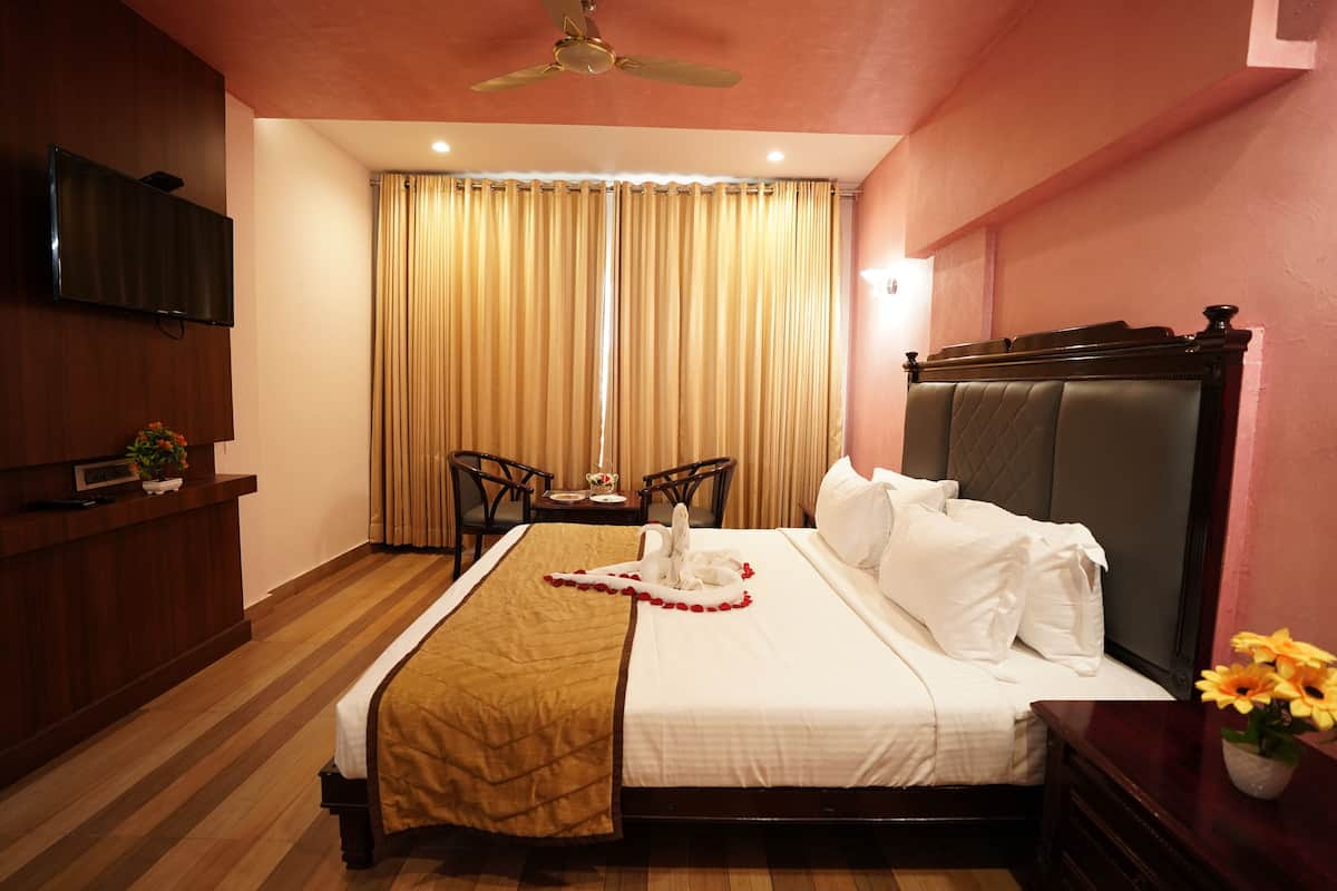 Une chambre d’hôtel avec un lit, une télévision, un ventilateur de plafond, une fenêtre avec des rideaux, une petite table sur laquelle est posé un vase de fleurs et un plancher en bois.