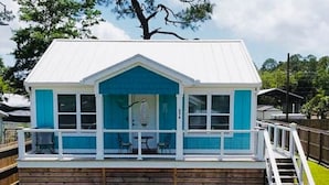 A small, single-story building with a white roof and blue trim, featuring a covered deck with a railing and a staircase leading up to it.