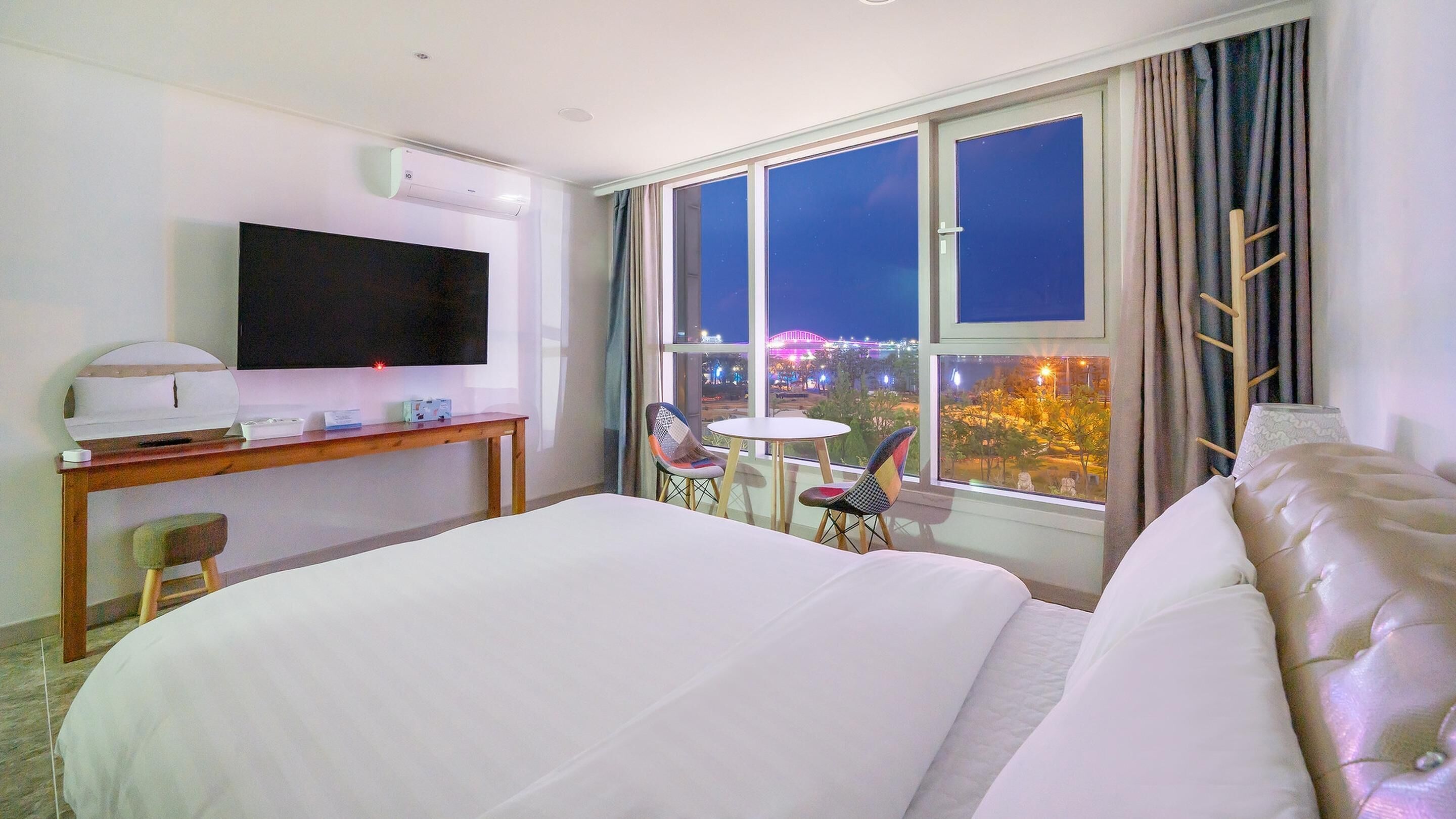 A hotel room with a large bed, a desk with a flat-screen TV, a small round table, two chairs, and a window with a city view at night.