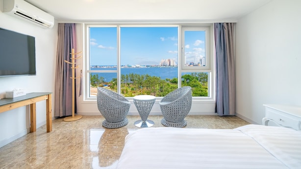 A hotel room with a large window offering a view of the sea and cityscape, a bed, a TV, and two patterned chairs. - Sokcho Ocean View Pension (Sokcho)