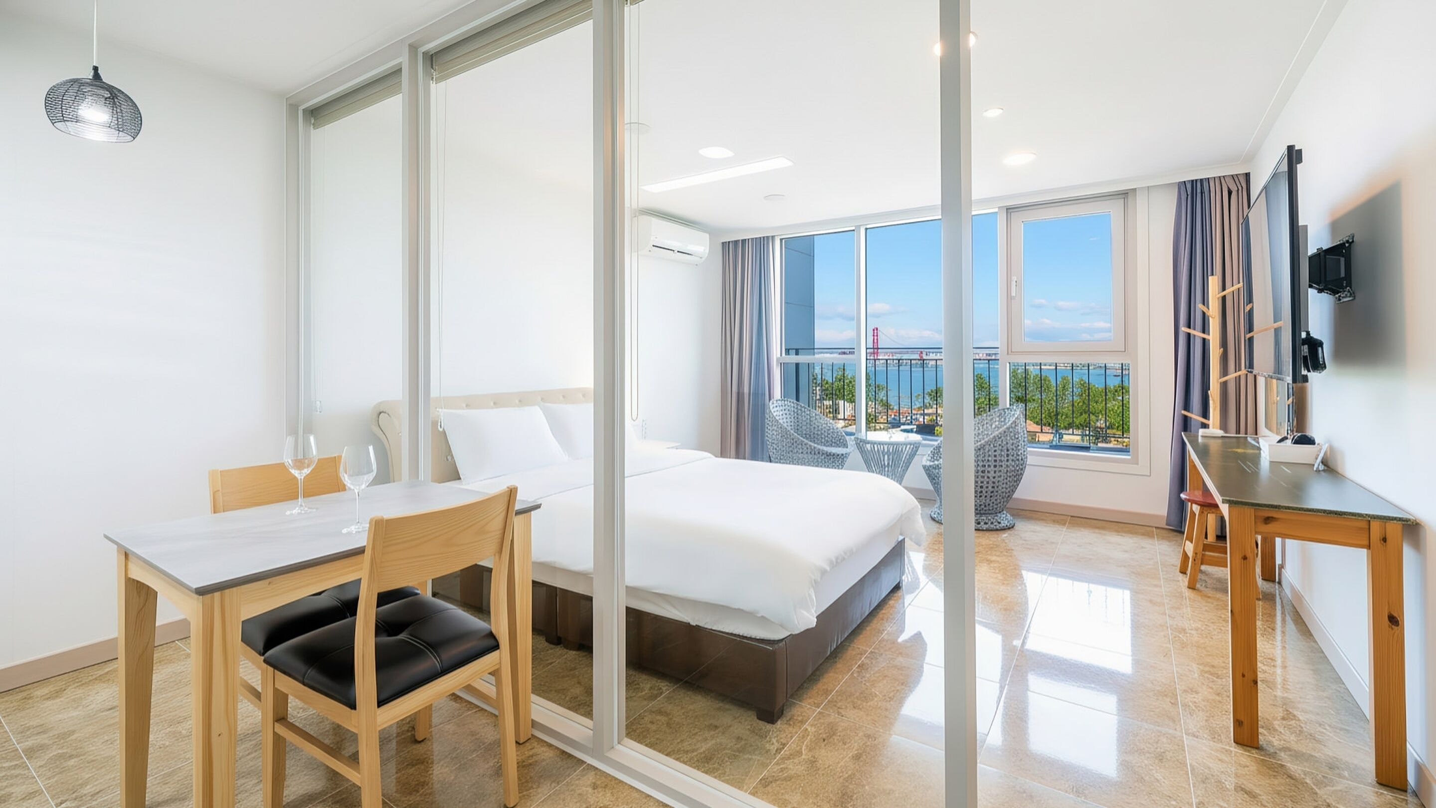 A modern hotel room with a bed, a dining area with a table and chairs, a TV mounted on the wall, and a large window offering a view of the sea.