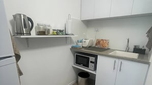 Pieni, käytännöllinen keittiö, jossa on valkoiset kaapit, mikroaaltouuni, vedenkeitin ja roskakori.