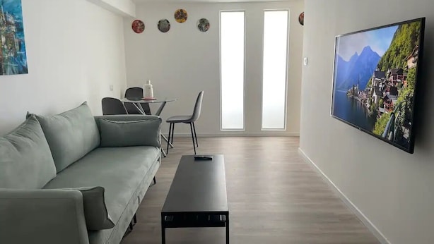 Moderni olohuone, jossa on harmaa sohva, sohvapöytä ja seinälle asennettu televisio. Huoneessa on kaksi ikkunaa, joissa on sälekaihtimet, ovi ja seinälle ripustettu taideteos.
