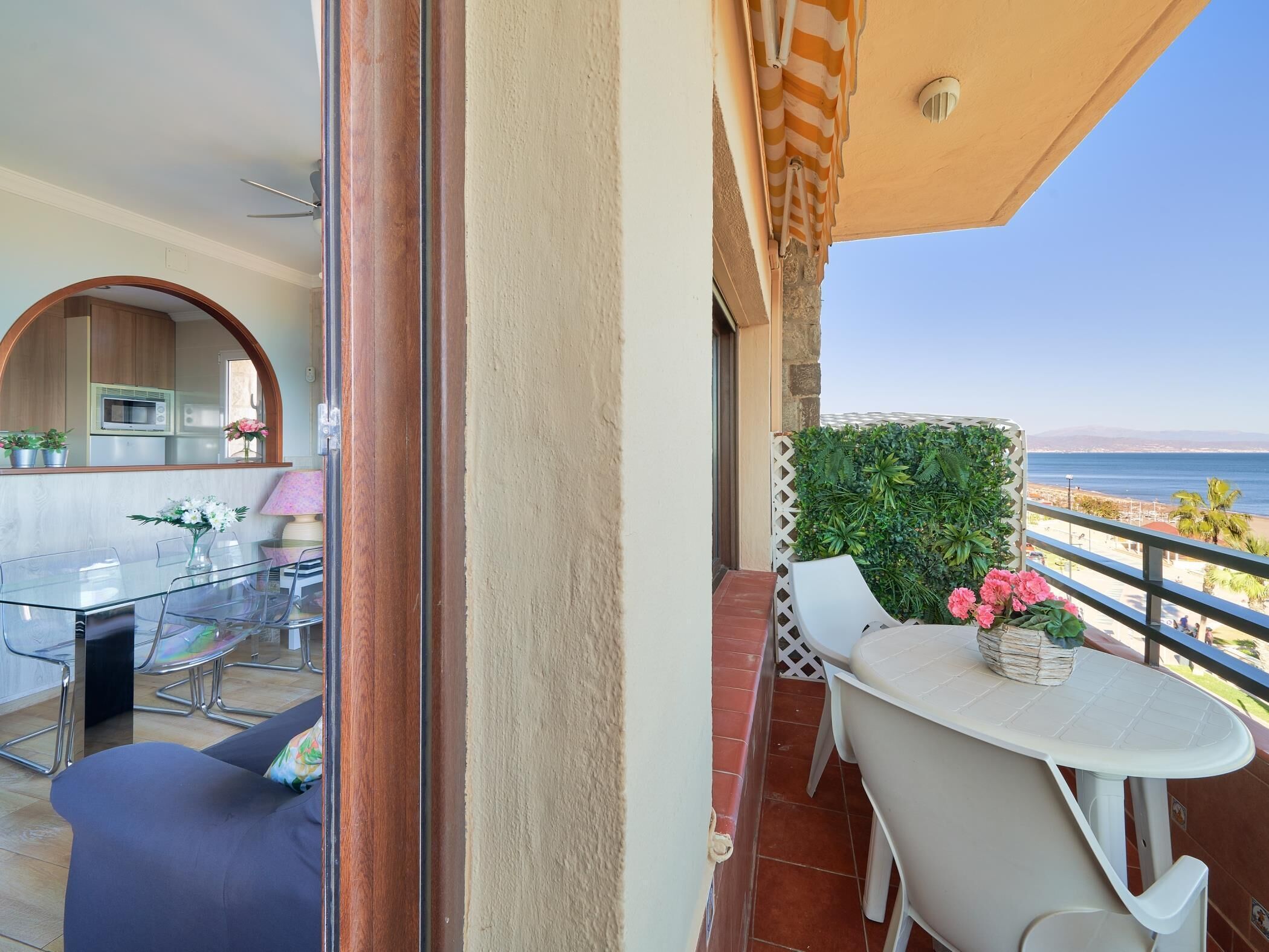 Un balcón con vistas al mar, una zona de comedor con mesa y sillas, y un pequeño rincón de estar con un cojín.
