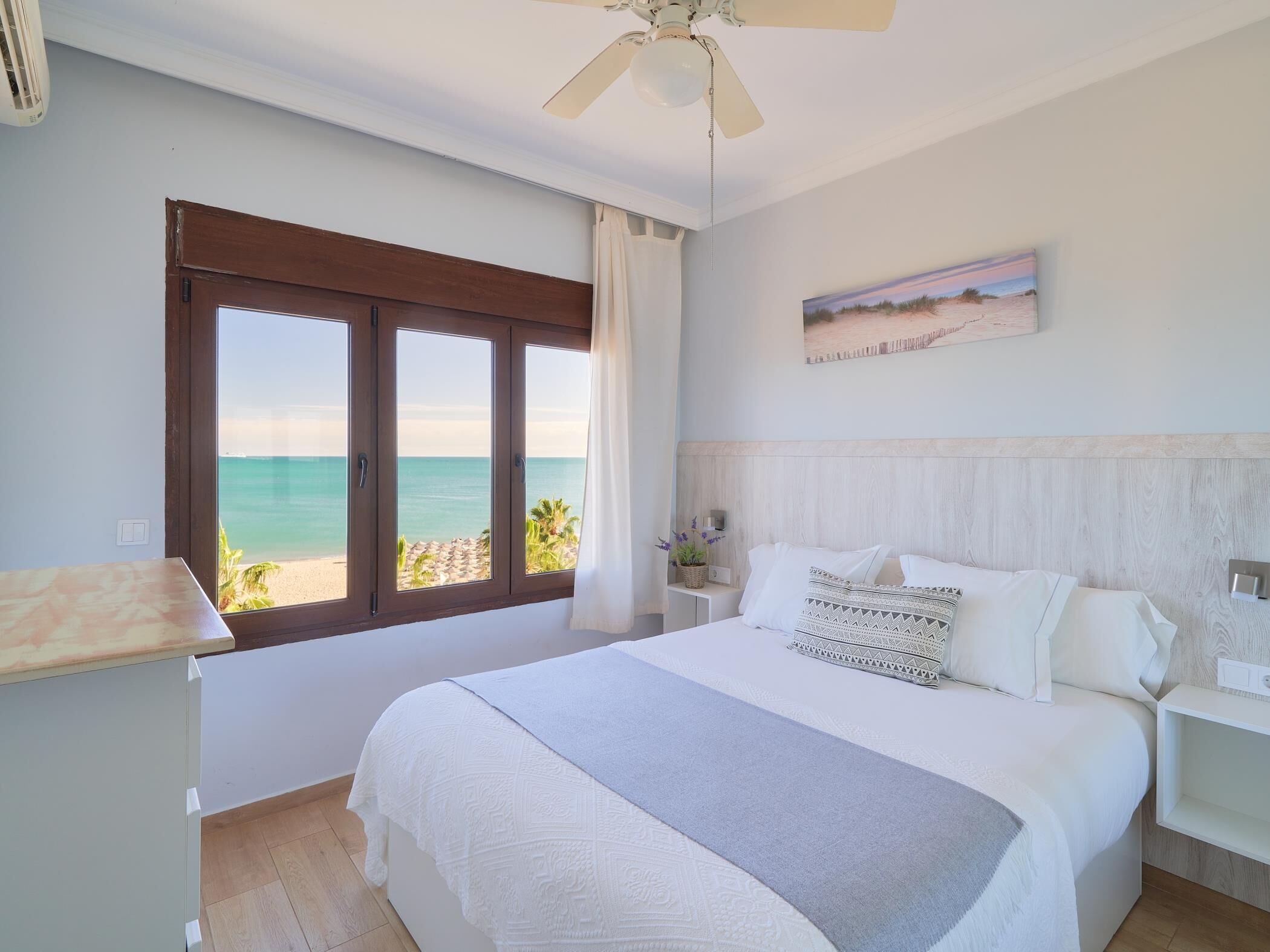 Un dormitorio con una cama grande, un ventilador de techo, vista a la playa y un cuadro en la pared.