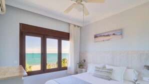 Une chambre avec un grand lit, un ventilateur de plafond, une vue sur la plage et un tableau au mur.