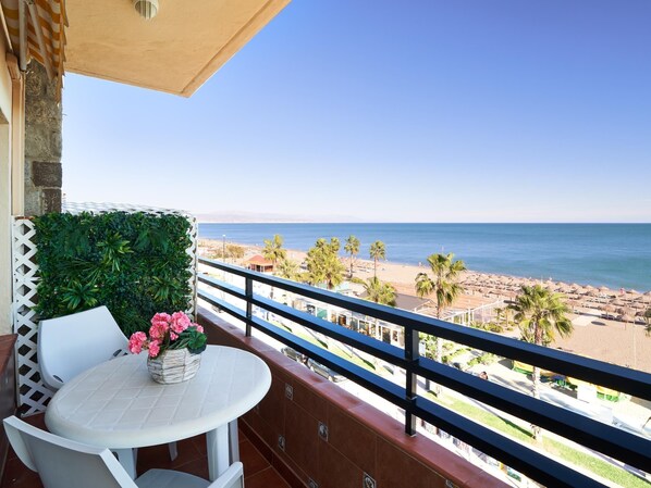 Un balcón con una mesa y sillas blancas, una planta en maceta y vistas a la playa y el océano.