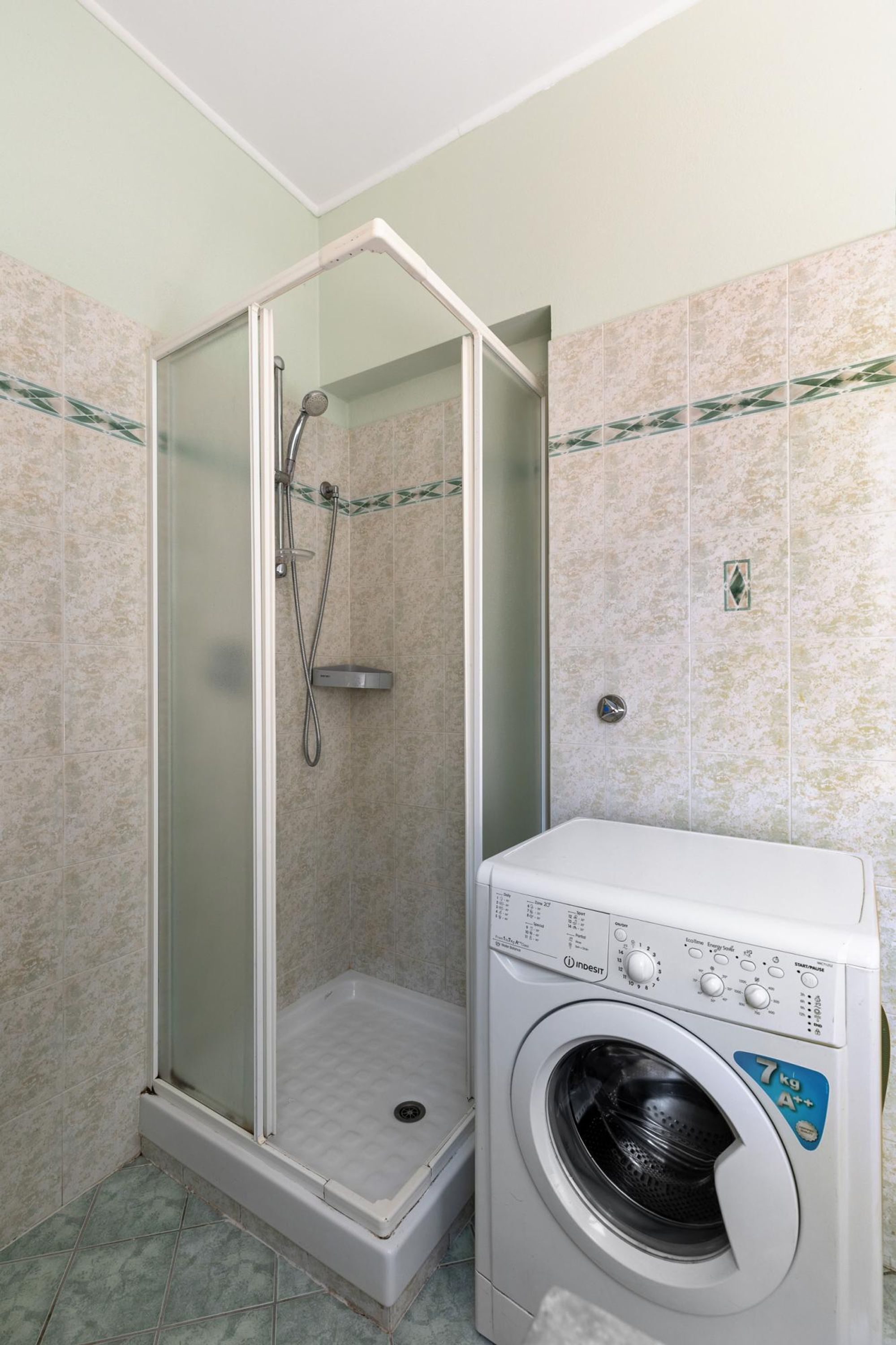 A compact bathroom with a glass-enclosed shower, a built-in washing machine, and tiled walls.