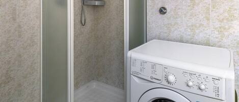 A compact bathroom with a glass-enclosed shower, a built-in washing machine, and tiled walls.