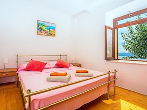 Un dormitorio con cama, almohadas, una mesita de noche, lámpara, ventana con vistas y un cuadro en la pared.