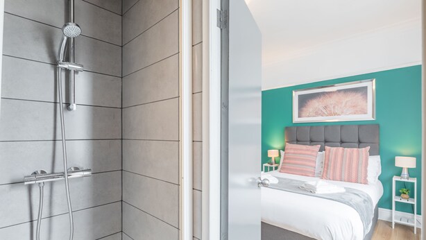 A modern bathroom with a shower and a bedroom with a bed, bedside tables, and a wall-mounted artwork.