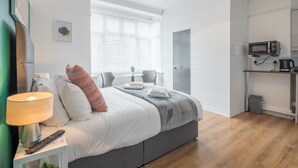 A bedroom with a bed, bedside table, lamp, and a view of a kitchen area.