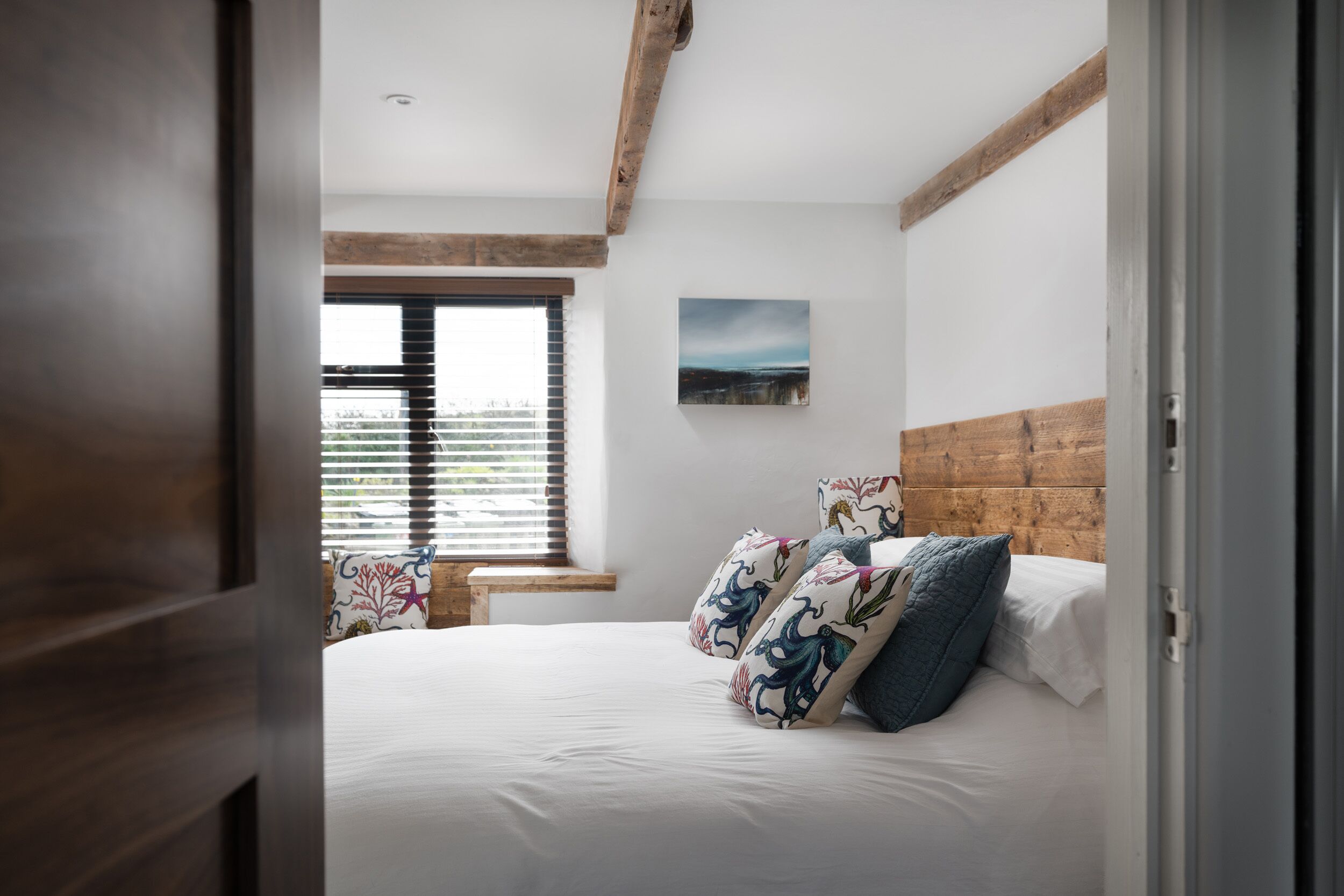 Une chambre avec une tête de lit en bois, un grand lit agrémenté d’oreillers à motifs, une fenêtre avec des stores et une petite table placée près de la fenêtre.