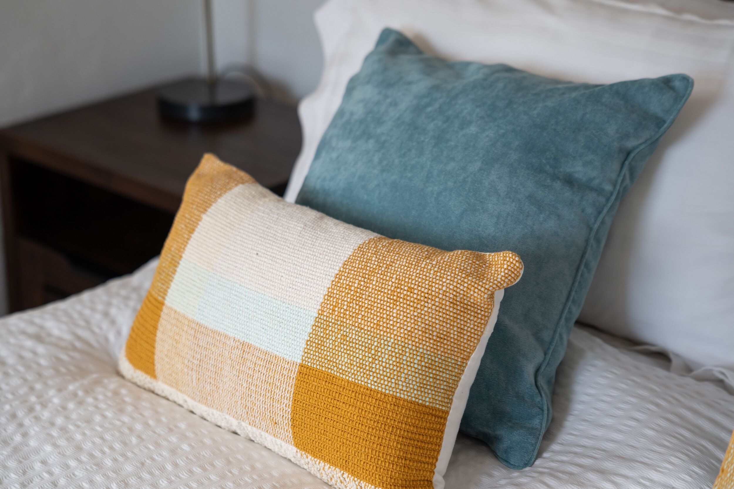 A bed with a white textured blanket, a mustard-colored pillow, and a teal pillow.