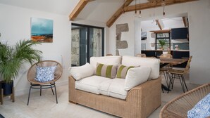 Ein gemütlicher Wohnbereich mit einem Rattan-Sofa, einem Esstisch und Stühlen. Ein großes Fenster bietet einen Blick ins Freie.