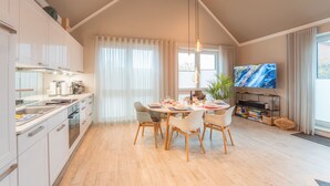 Et moderne køkken med lyse skabe, et spiseområde med et rundt bord og fire stole, et fladskærms-tv og et stort vindue med gardiner.