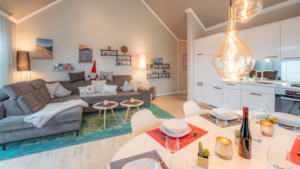 En moderne stue med en grå sofa, et spisebord dækket til to personer, og et køkken med hvide skabe og en komfur.