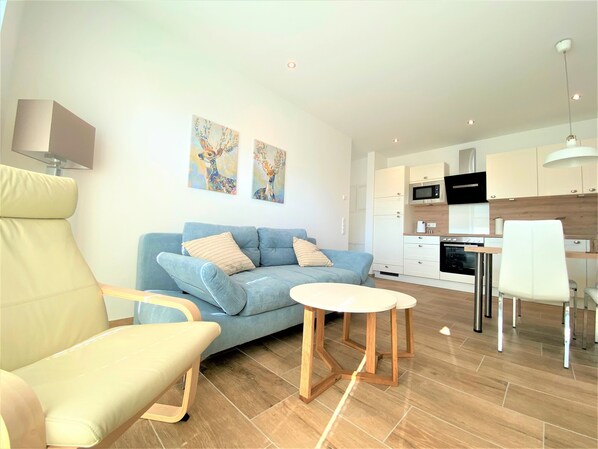 A modern living room with a blue sofa, a wooden coffee table, and a beige armchair. There is a kitchen area with white cabinets and a microwave. - Hello and welcome to Apartment Fiete! (Dangast)