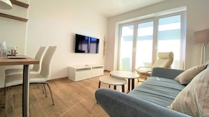 Un salon moderne comprenant un canapé, une table à manger avec des chaises, un téléviseur à écran plat et une grande fenêtre avec des stores.