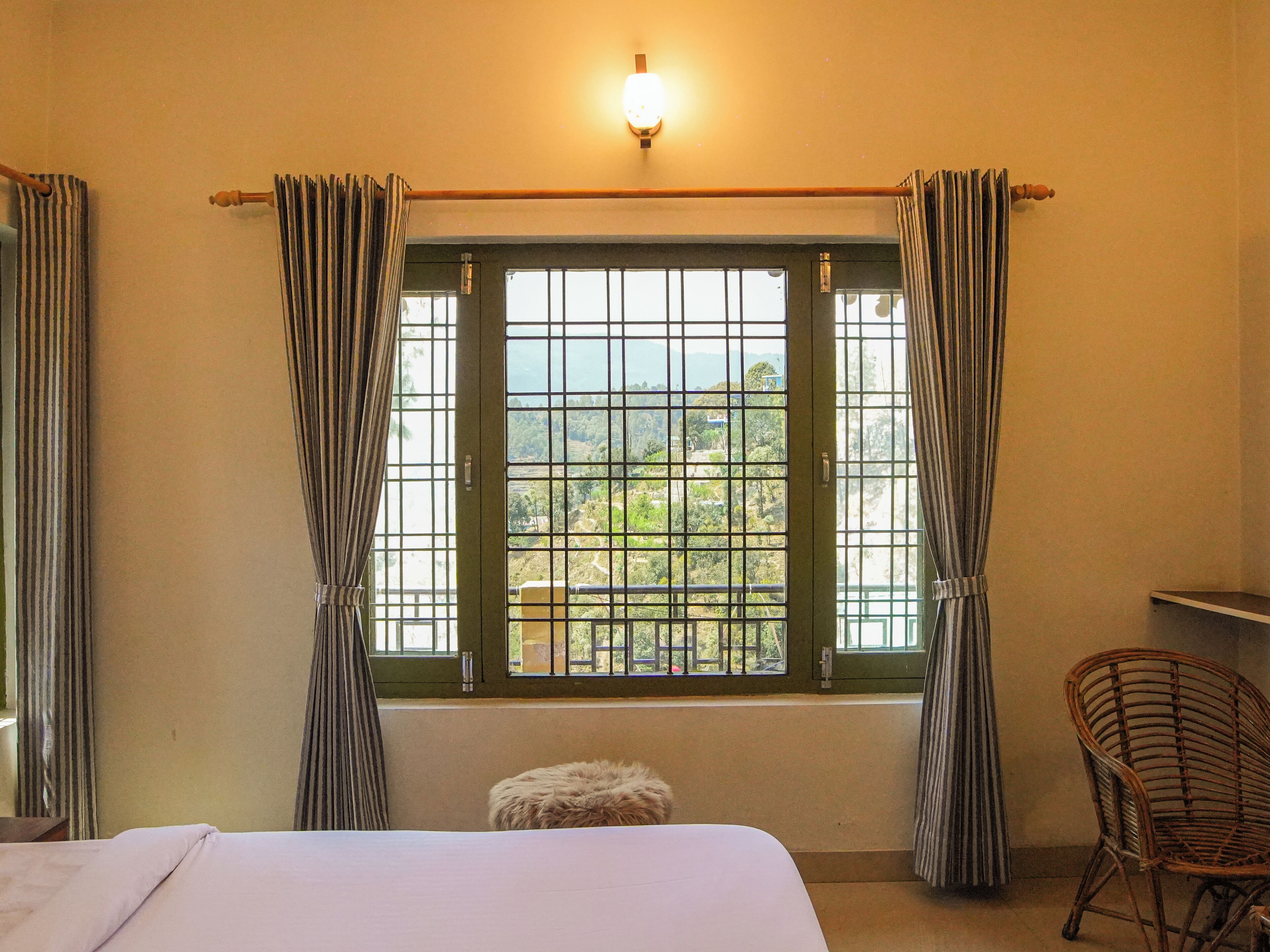 A room with a bed, a chair, a window with curtains, and a view of greenery outside.