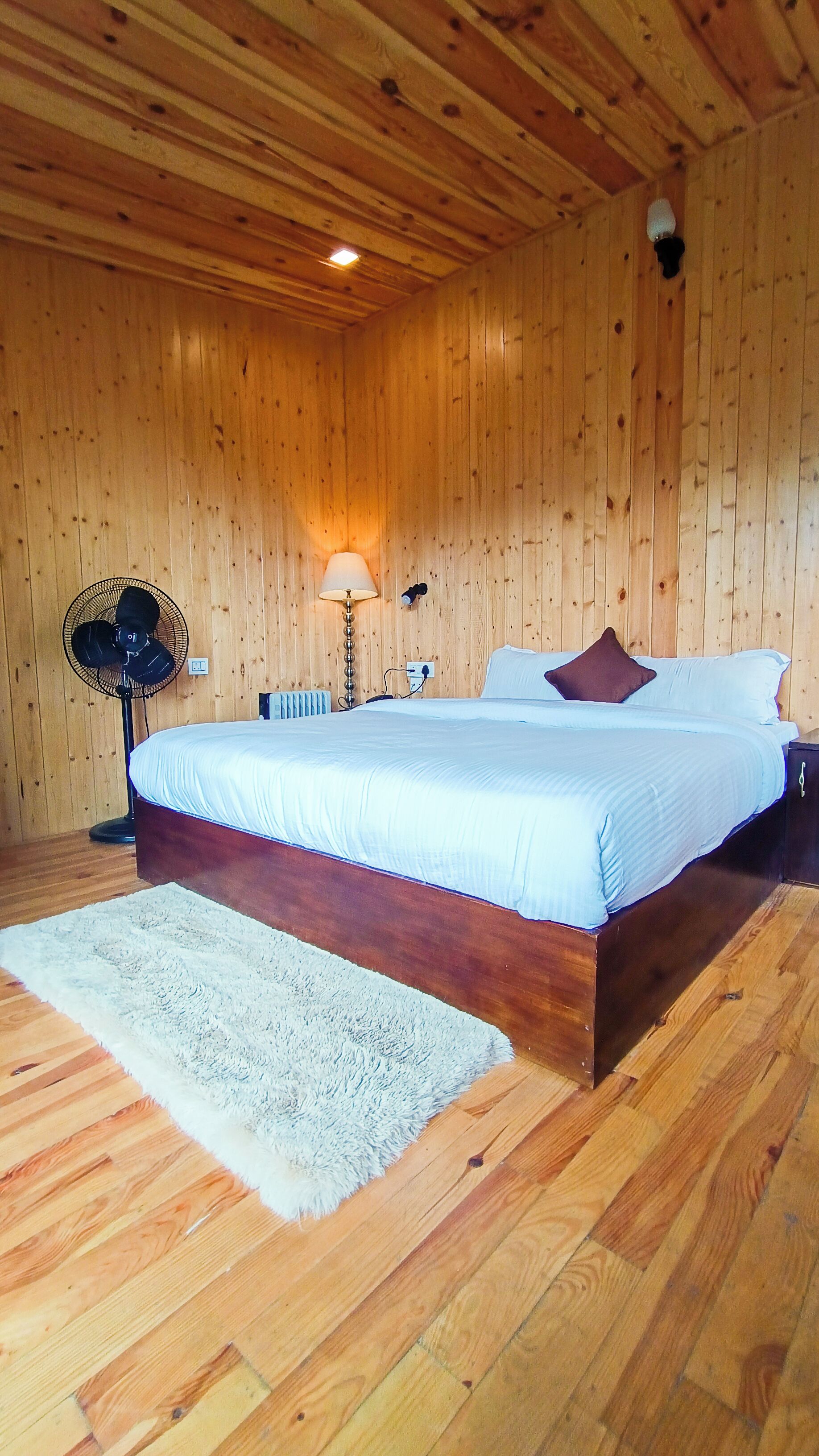 A wooden-paneled room with a bed, a standing fan, a lamp, and a white rug.