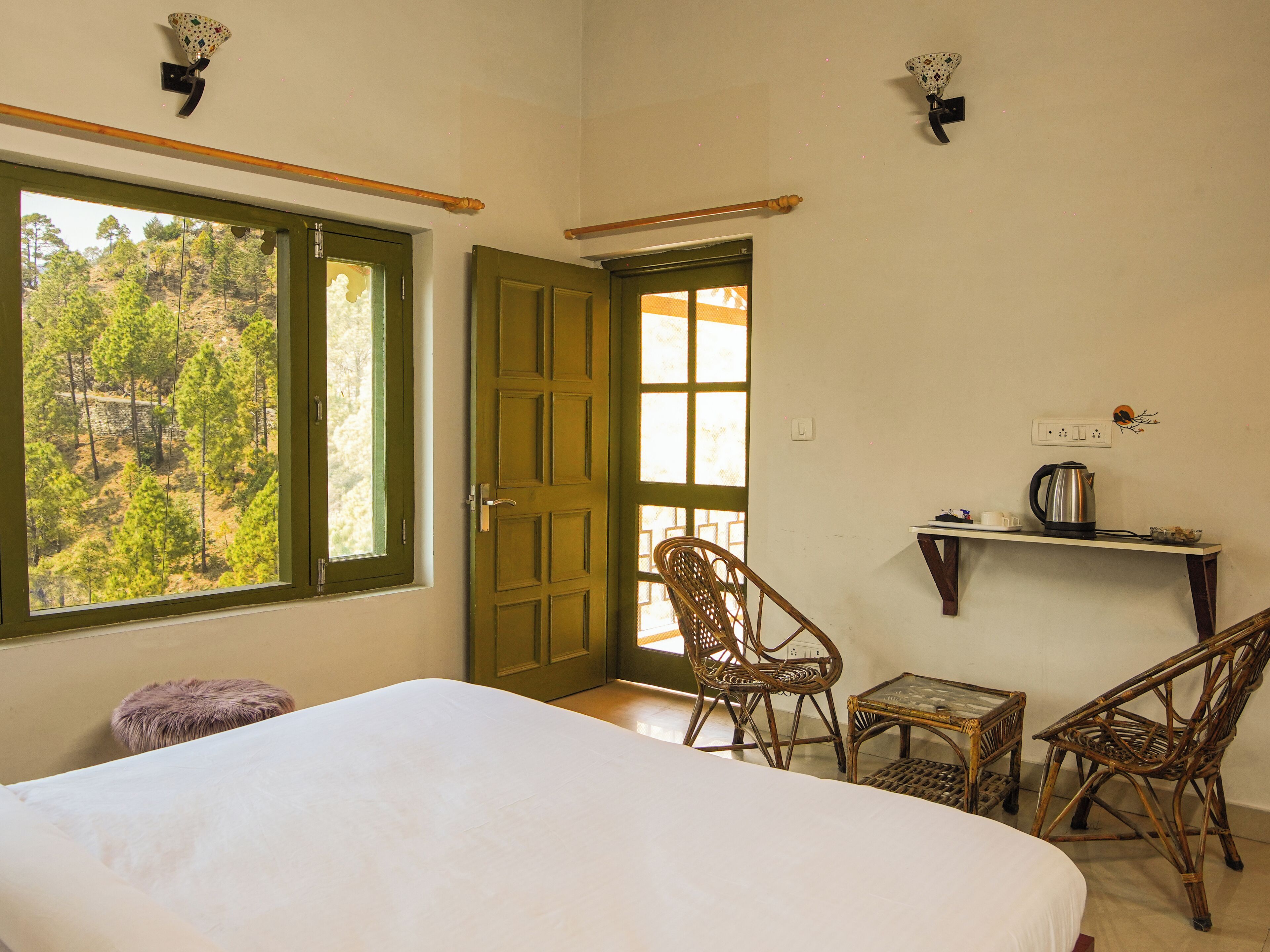 A room with a bed, two wicker chairs, a small table, a window with a view of trees, and a door.