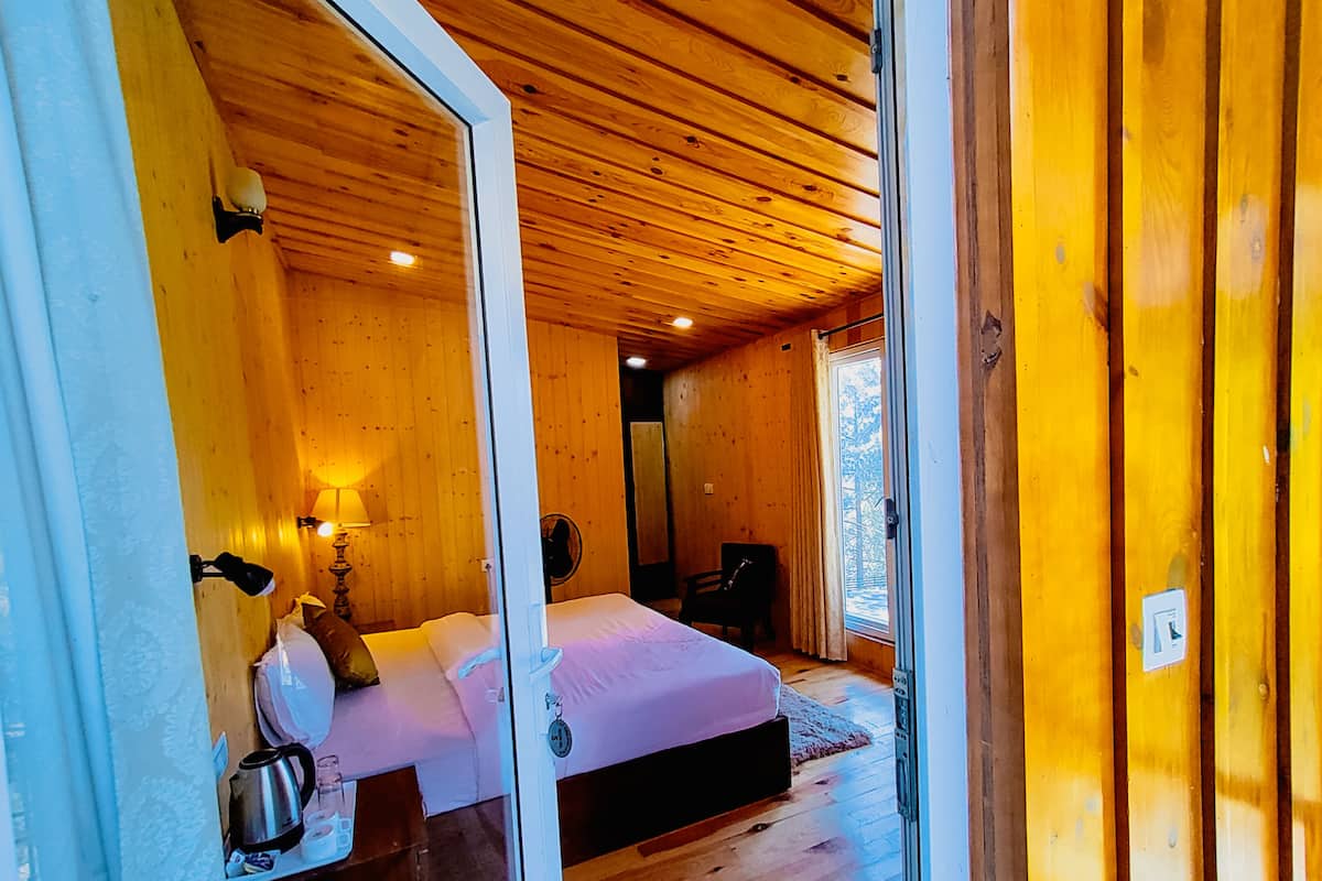Un dormitorio de cabaña de madera con cama, mesita de noche, lámpara y vistas al exterior.