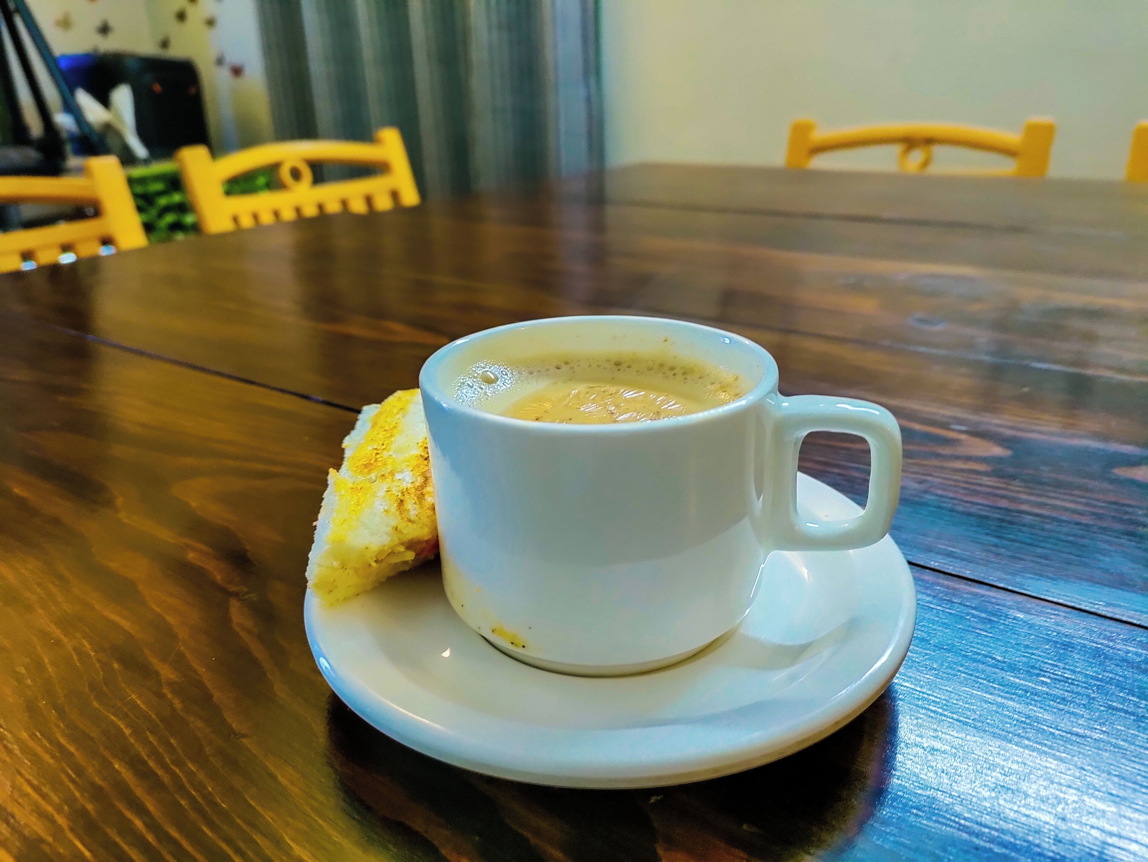 Una taza de café blanco sobre un platillo, con una tostada sobre la mesa.