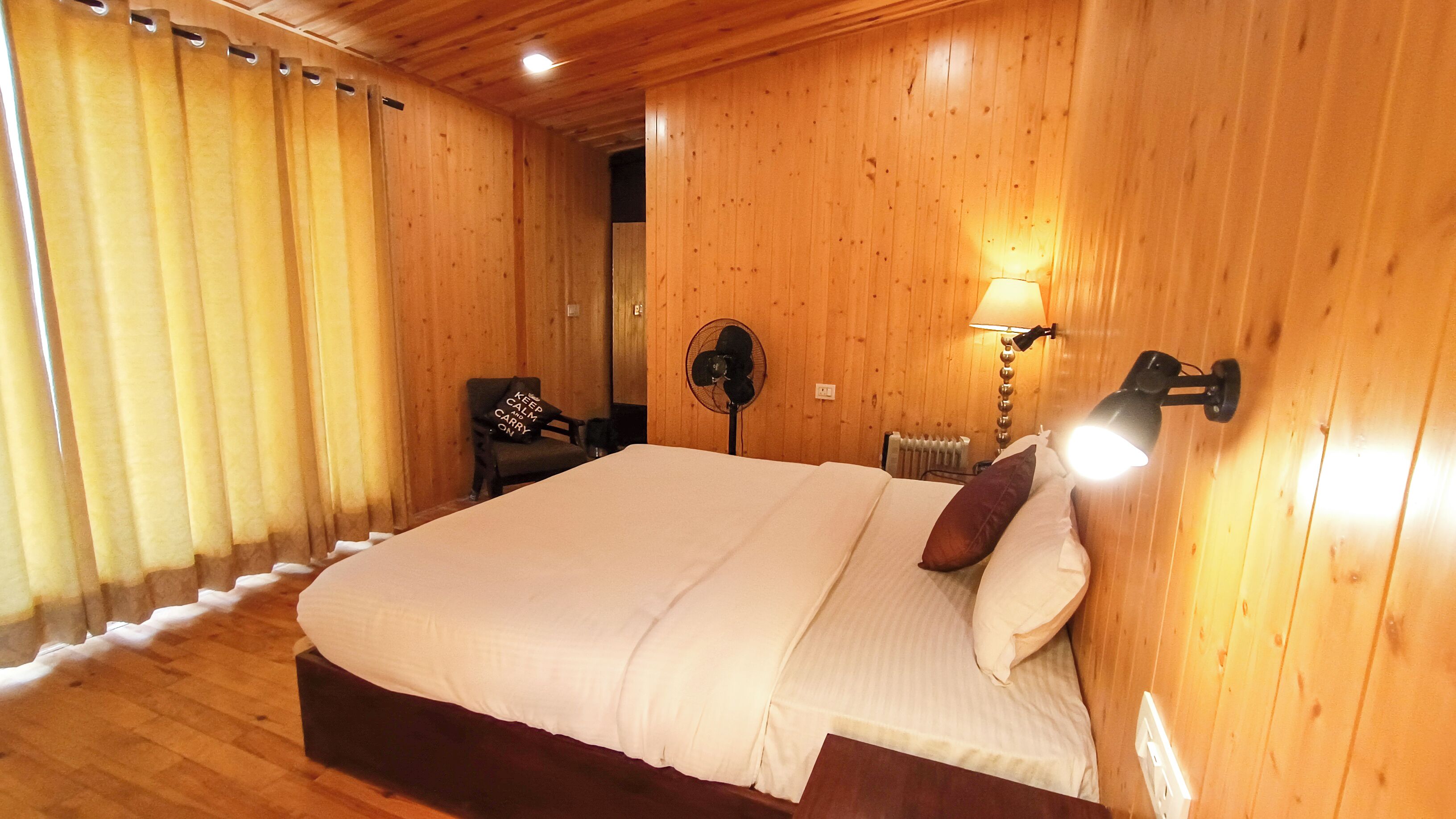 A wooden-paneled bedroom with a large bed, a standing fan, a chair, and a bedside lamp.