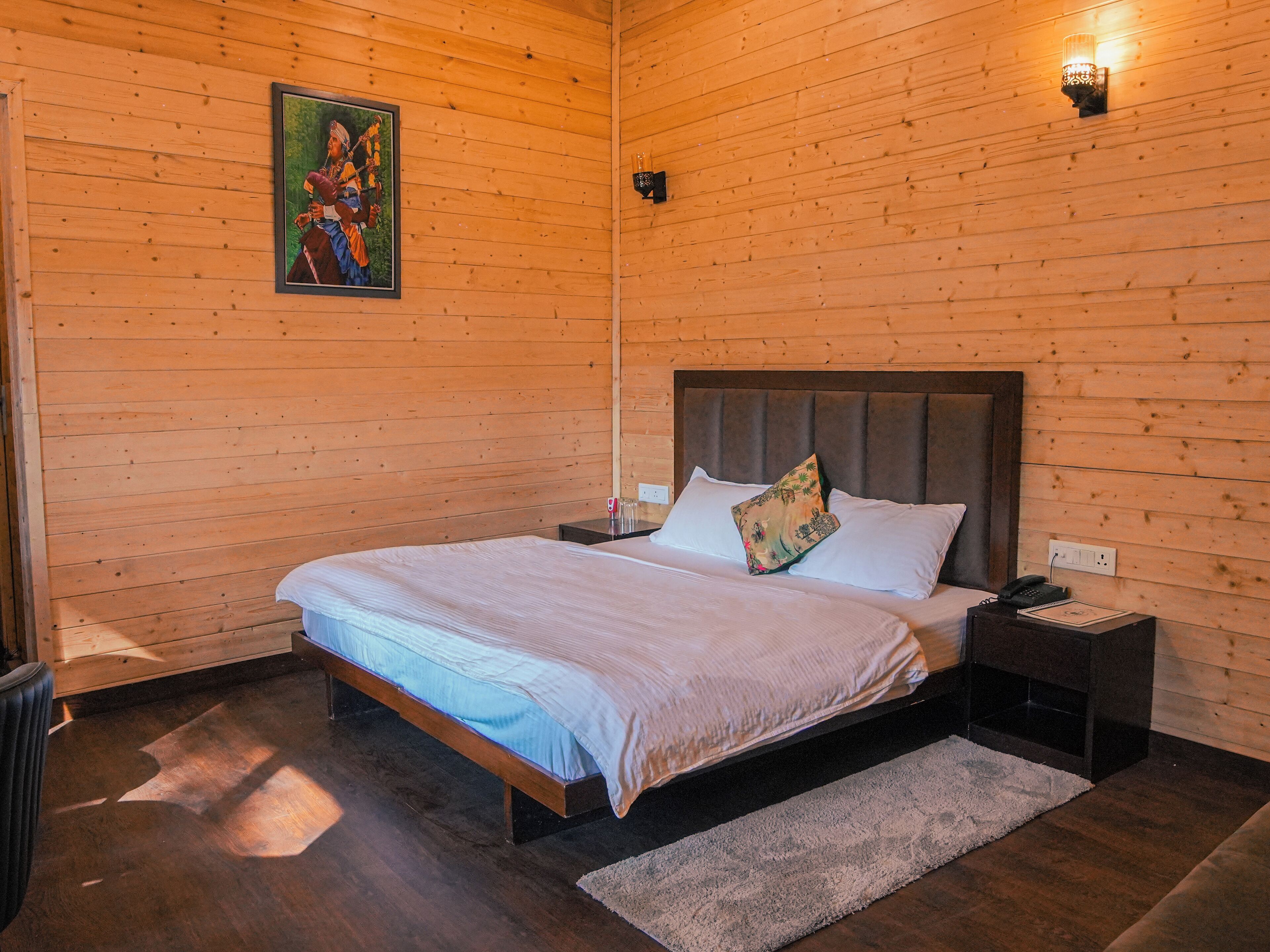 Una habitación con paneles de madera, una cama, una mesita de noche, una lámpara de pared y un cuadro enmarcado.