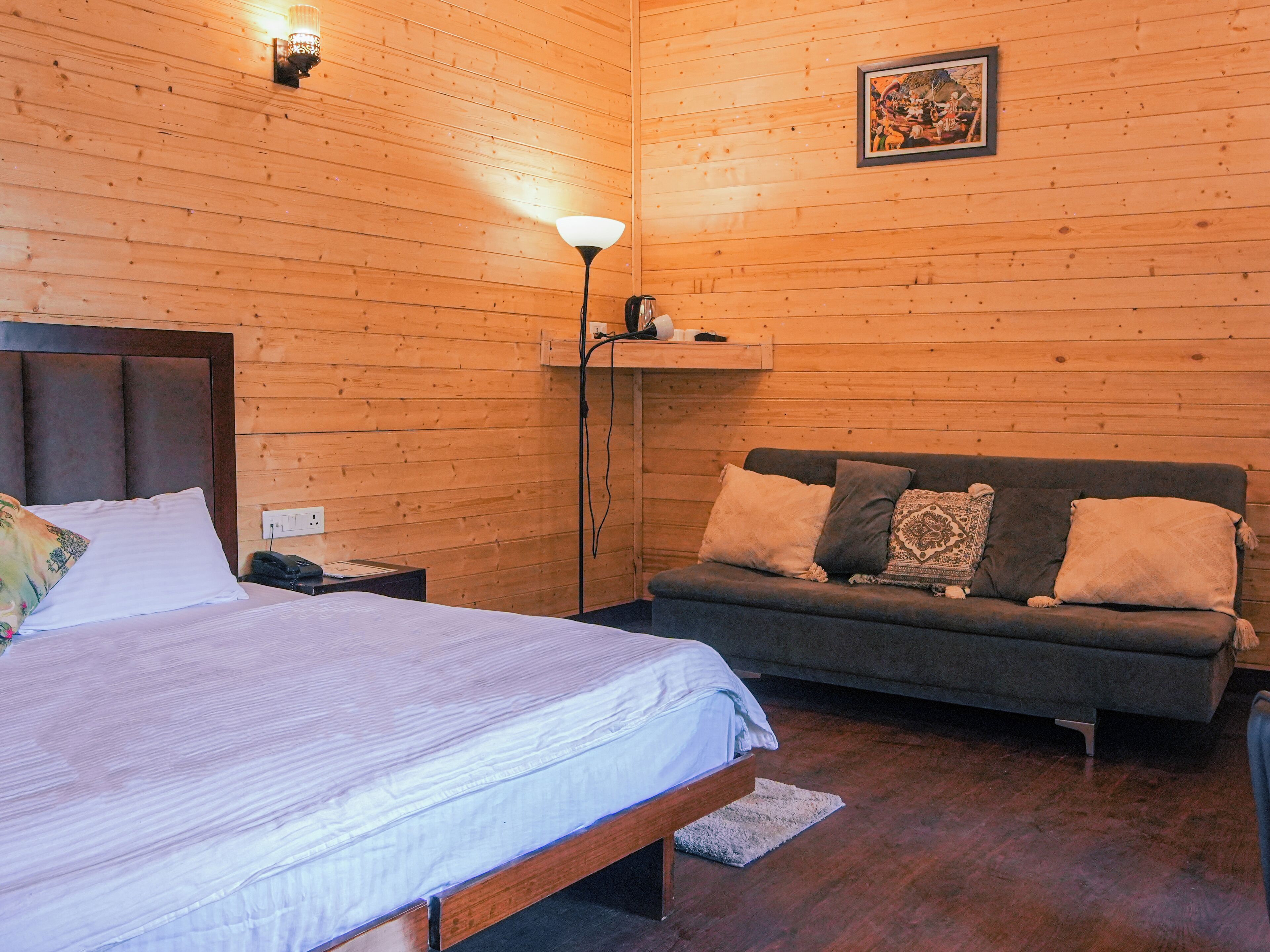 A wooden-paneled room with a bed, a sofa, a lamp, and a framed picture on the wall.