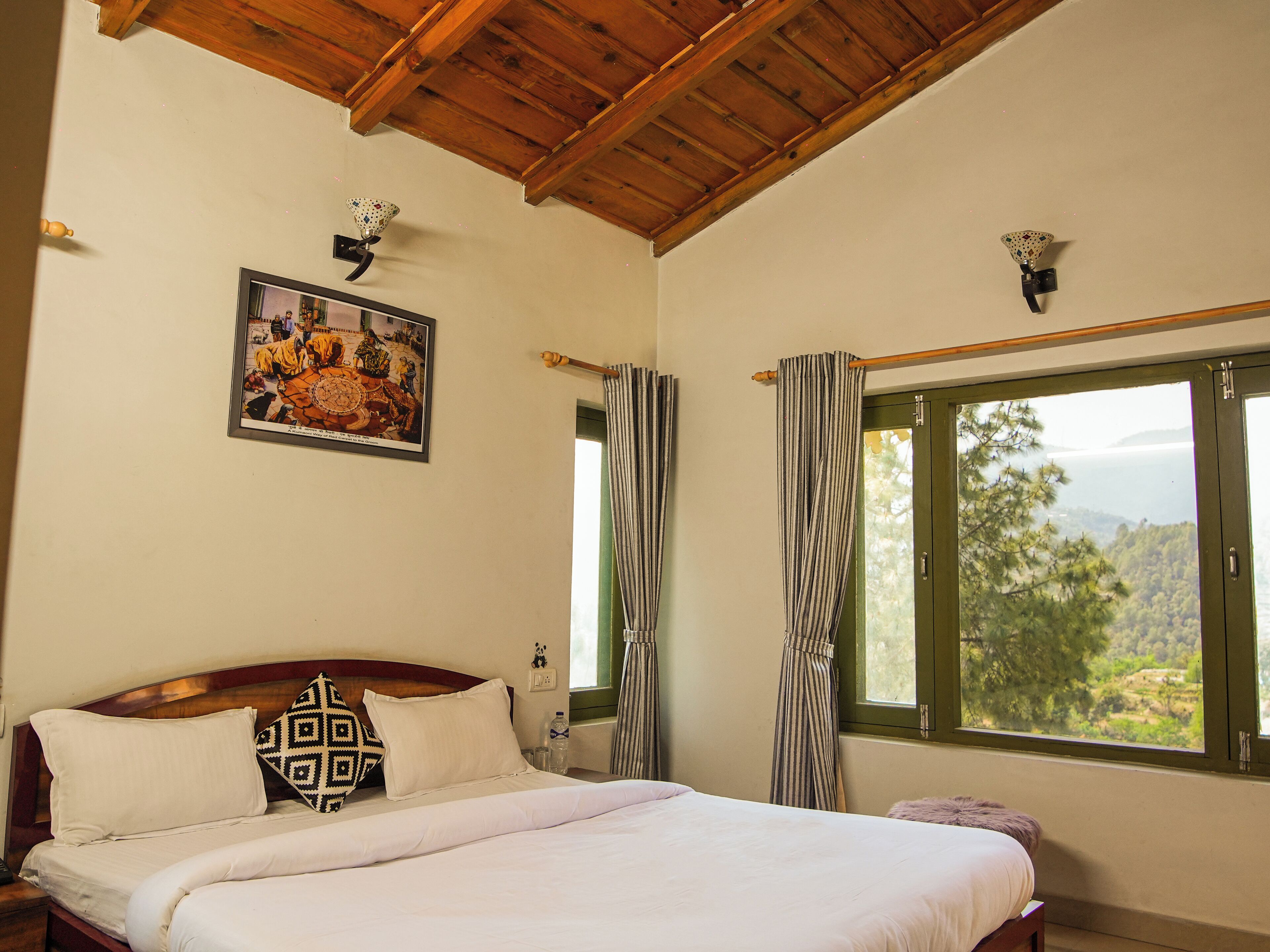 A bedroom with a wooden ceiling, a bed with white bedding, striped curtains, and a window with a view of trees and hills.