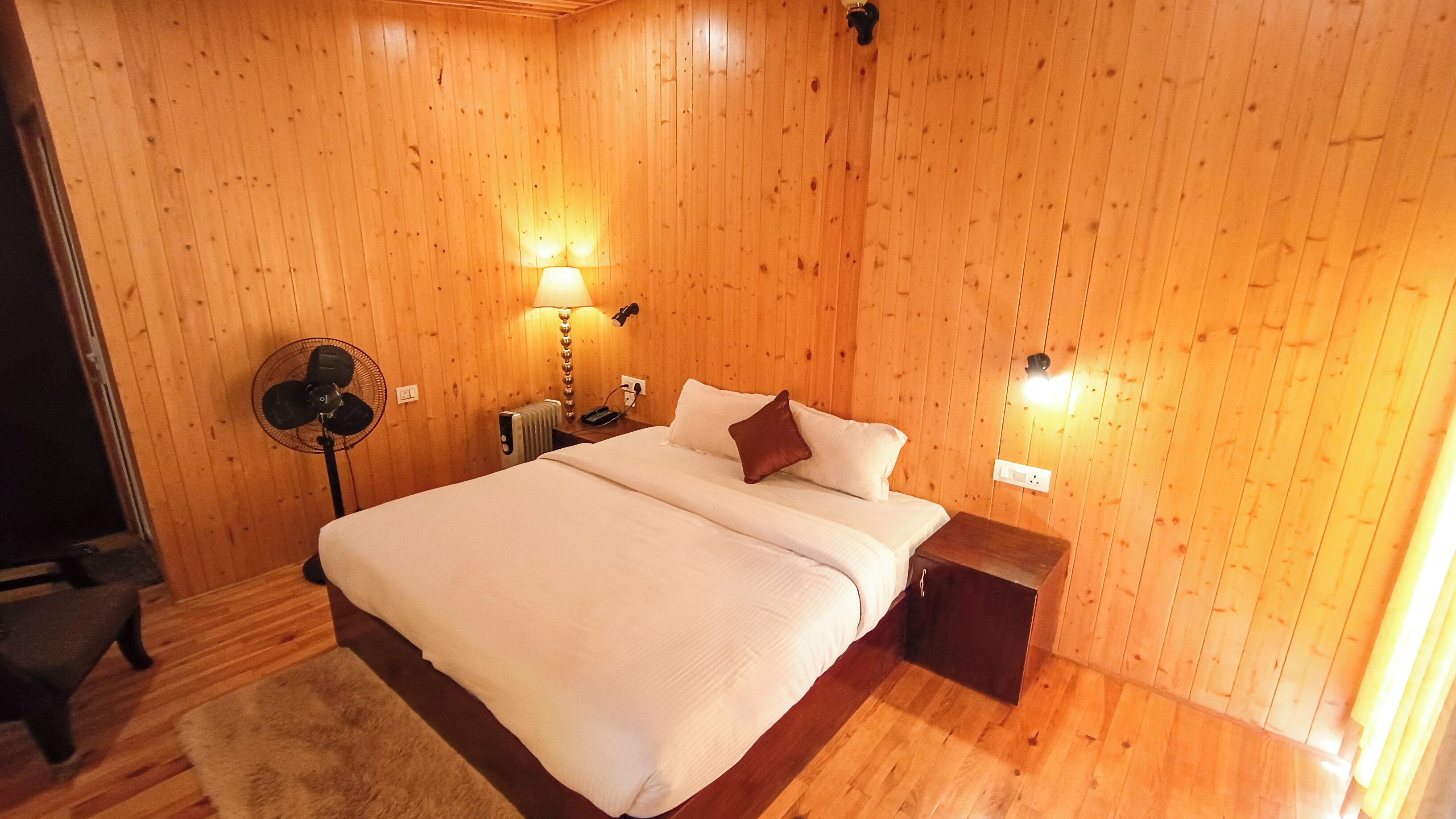 Habitación con paneles de madera, una cama, una mesita de noche, una lámpara y un ventilador.